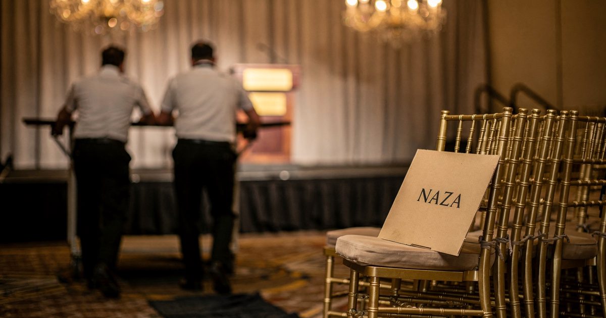 Under warm chandeliers in a hotel ballroom during late-afternoon setup, two catering staff seen from behind push a round banquet table away from the stage.