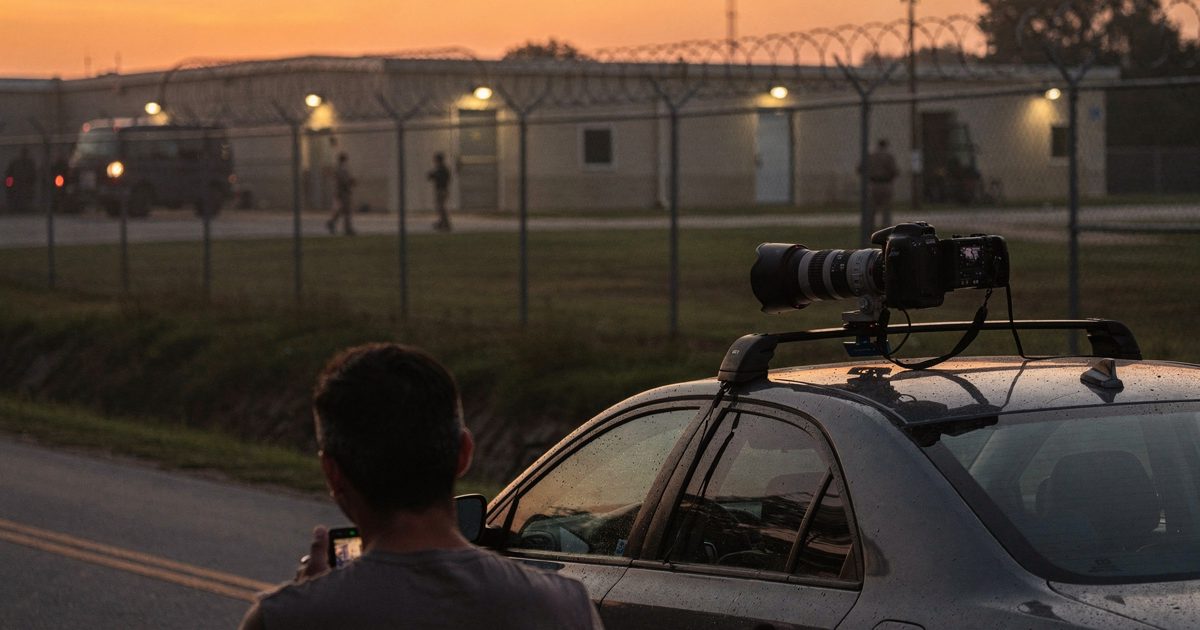 Humid golden dusk on a rural Louisiana roadside as a paparazzo cropped at the shoulders steadies a long telephoto lens across a rental car roof, a razor-wire.