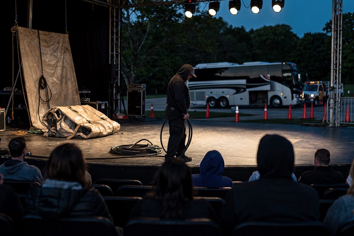 Shot from behind a small amphitheatre crowd at dusk, a lone stagehand coiling a cable under bare work lights on an unlit stage, a folded canvas backdrop.