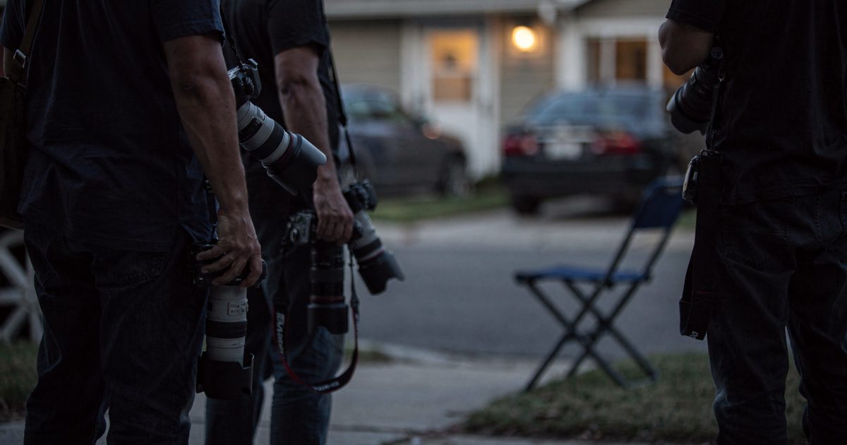 Photographers seen from behind on a residential sidewalk at dusk, long lenses lowered to their hips instead of raised, a folding camp stool left standing on.