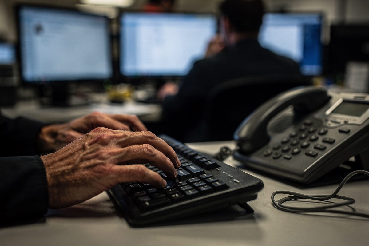 Tight close-up of a writer's hands resting still on a keyboard in a dim open-plan newsroom lit only by monitor glow, a desk phone off its cradle beside them.