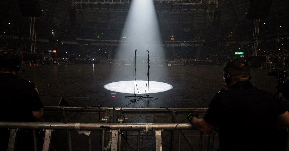 A view from behind the barricade of a massive stadium stage at night, where the blinding glare of a solitary follow-spot illuminates two microphone stands.