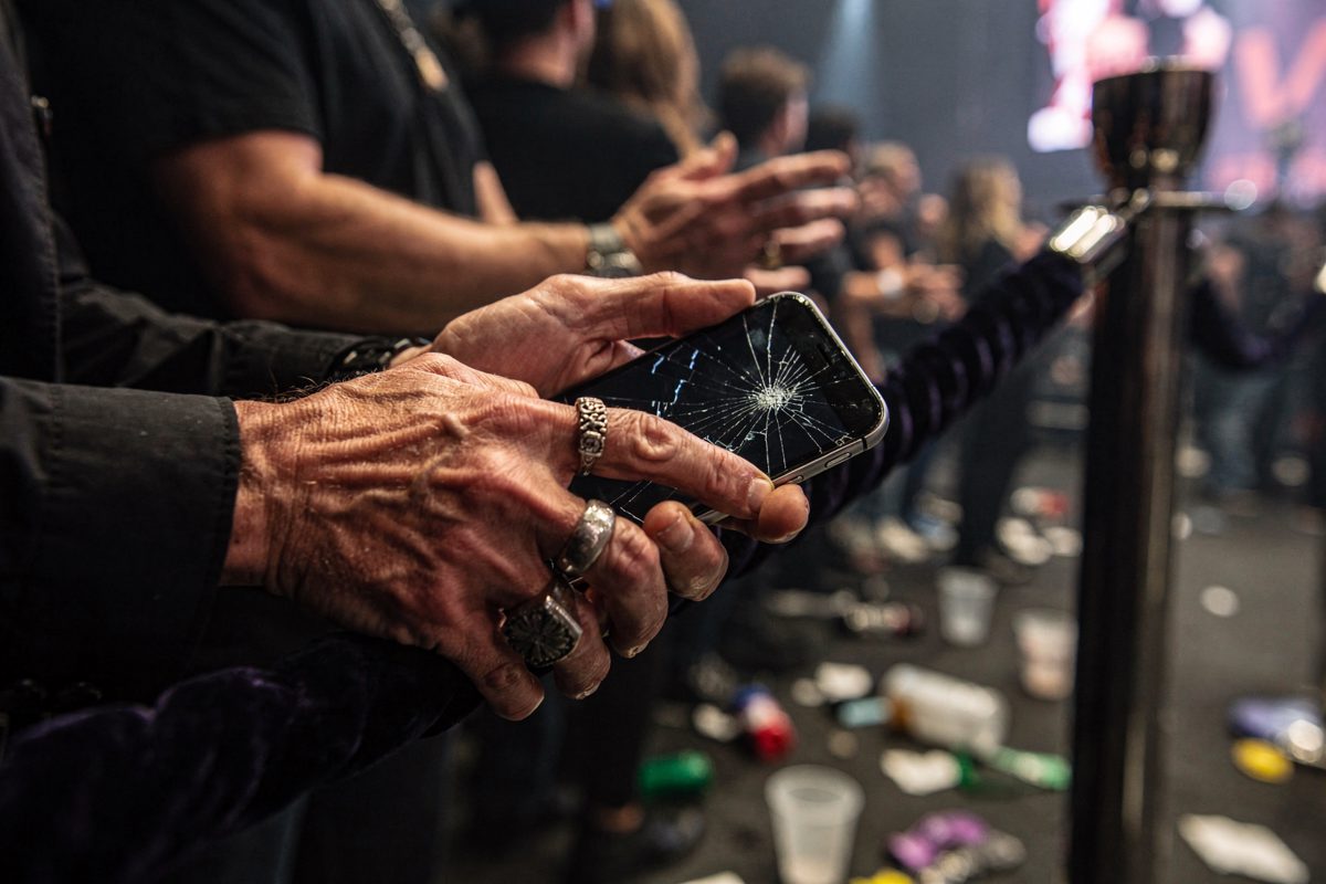 A frantic music executive's hands, heavily ringed and clutching a shattered smartphone, resting on the velvet rope of an exclusive VIP stadium enclosure.