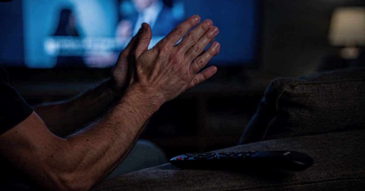 Close-up of two hands frozen mid-clap in the blue glow of a paused television in a dark living room late at night, a remote control abandoned face-down on the.