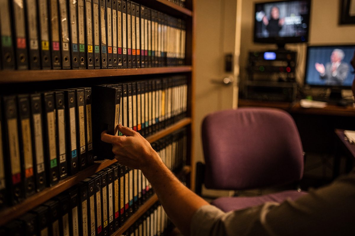 Behind-the-shoulder shot inside a production archive room in warm afternoon light, a part-cropped figure pulling a labeled videotape from a shelf stacked with.