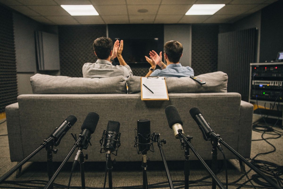 Wide shot of a soundproofed testing lab under flat fluorescent light, a row of microphones on boom stands aimed at a household sofa where two seated figures.