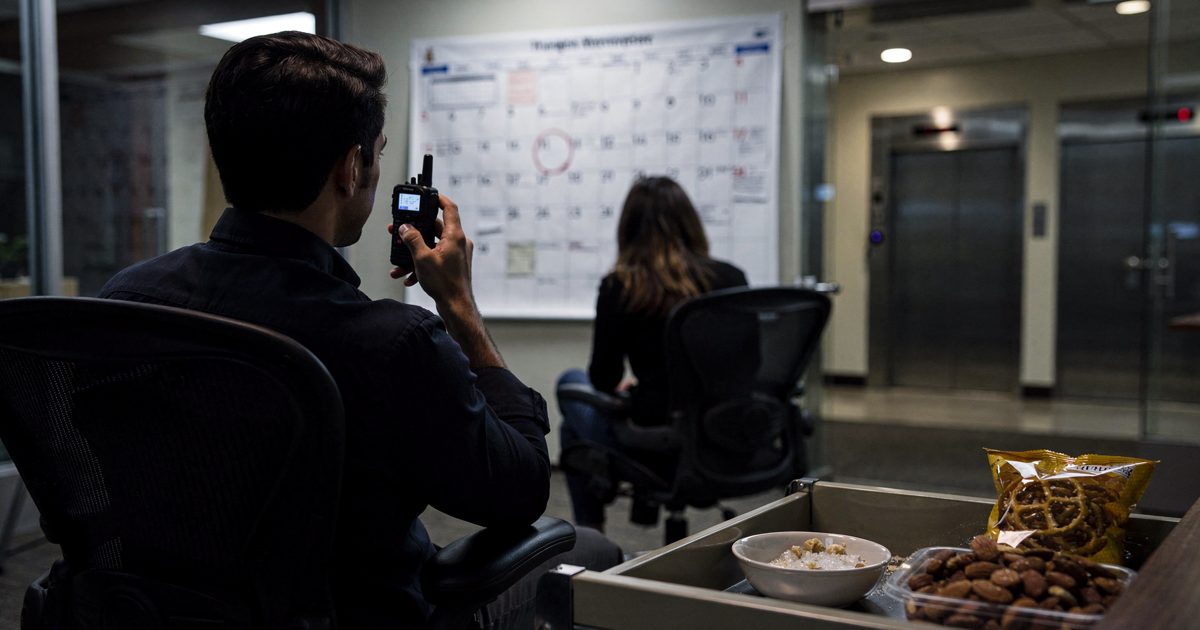 In a glass-walled office beside elevator banks after dark, two colleagues sit in rolling chairs facing a wall calendar while one, seen from behind, speaks into.