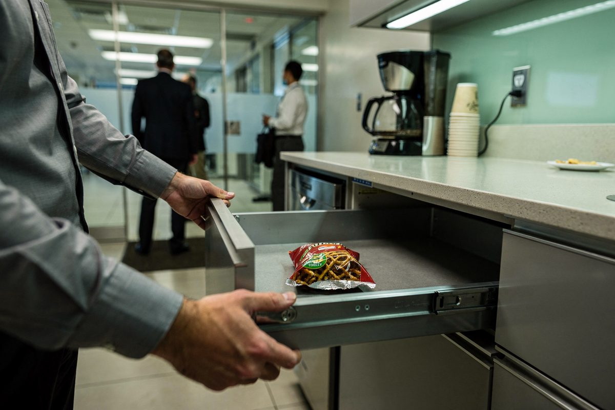 Close on a pair of hands in a corporate kitchenette under harsh morning fluorescents, pulling open an empty metal snack drawer with a crumpled pretzel bag on.