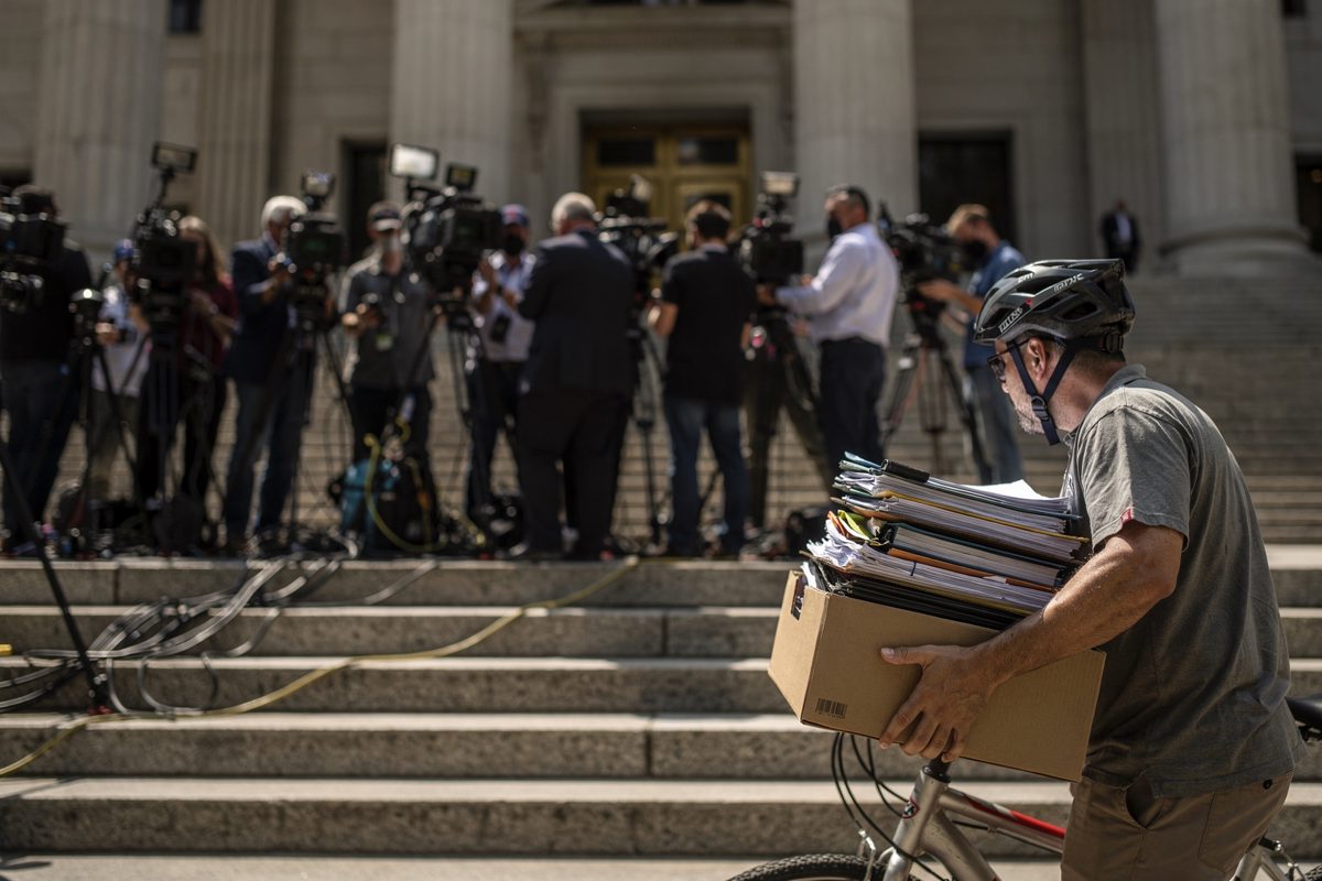 Wide shot at harsh midday on the granite steps outside the Manhattan criminal courthouse, a wall of television crews with their backs to the camera facing the.