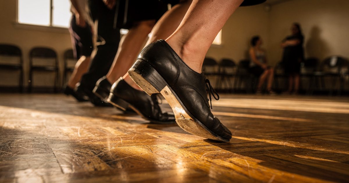 Low floor-level close detail in golden early-morning light across the scuffed parquet of a suburban church hall, a row of black tap shoes caught mid-step, one.