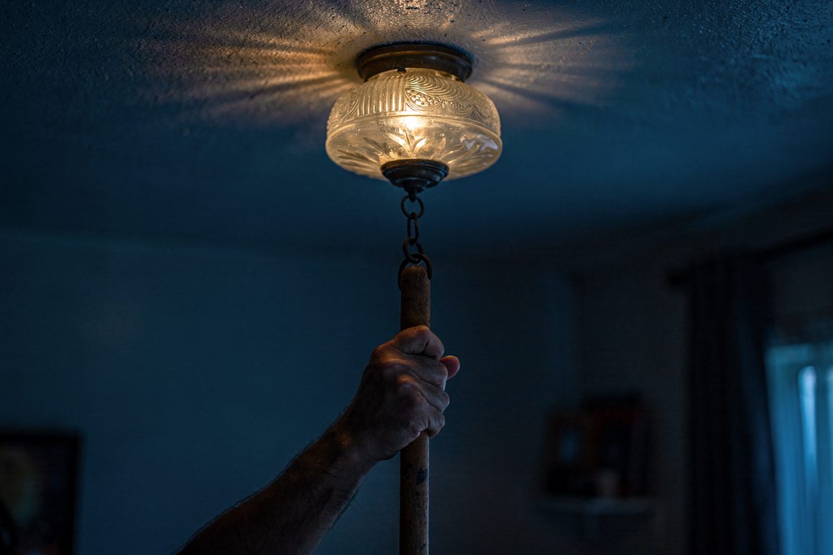 Close detail at blue dusk inside a downstairs apartment, a glass ceiling light fixture trembling on its chain while a hand cropped at the wrist raises a broom.