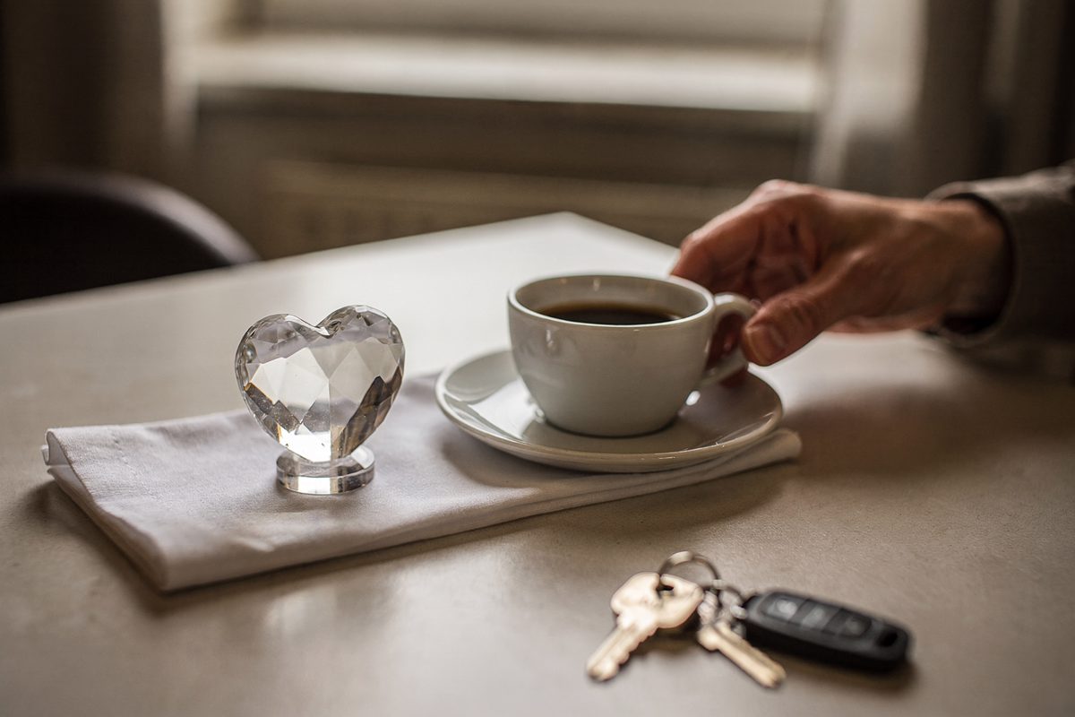 Morning window light on a hotel breakfast table, a small crystal heart standing beside an espresso cup on a folded napkin, one part-cropped hand reaching for.