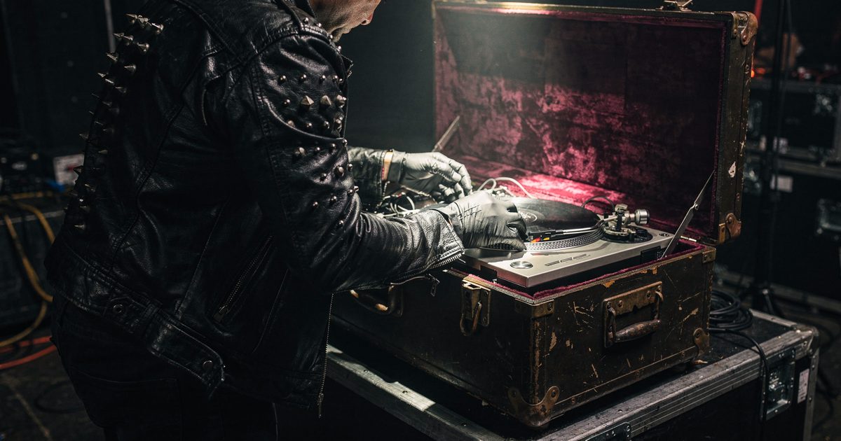 A close-up shot from behind of someone in a studded black leather jacket packing a pristine silver turntable into a velvet-lined vintage steamer trunk under a.