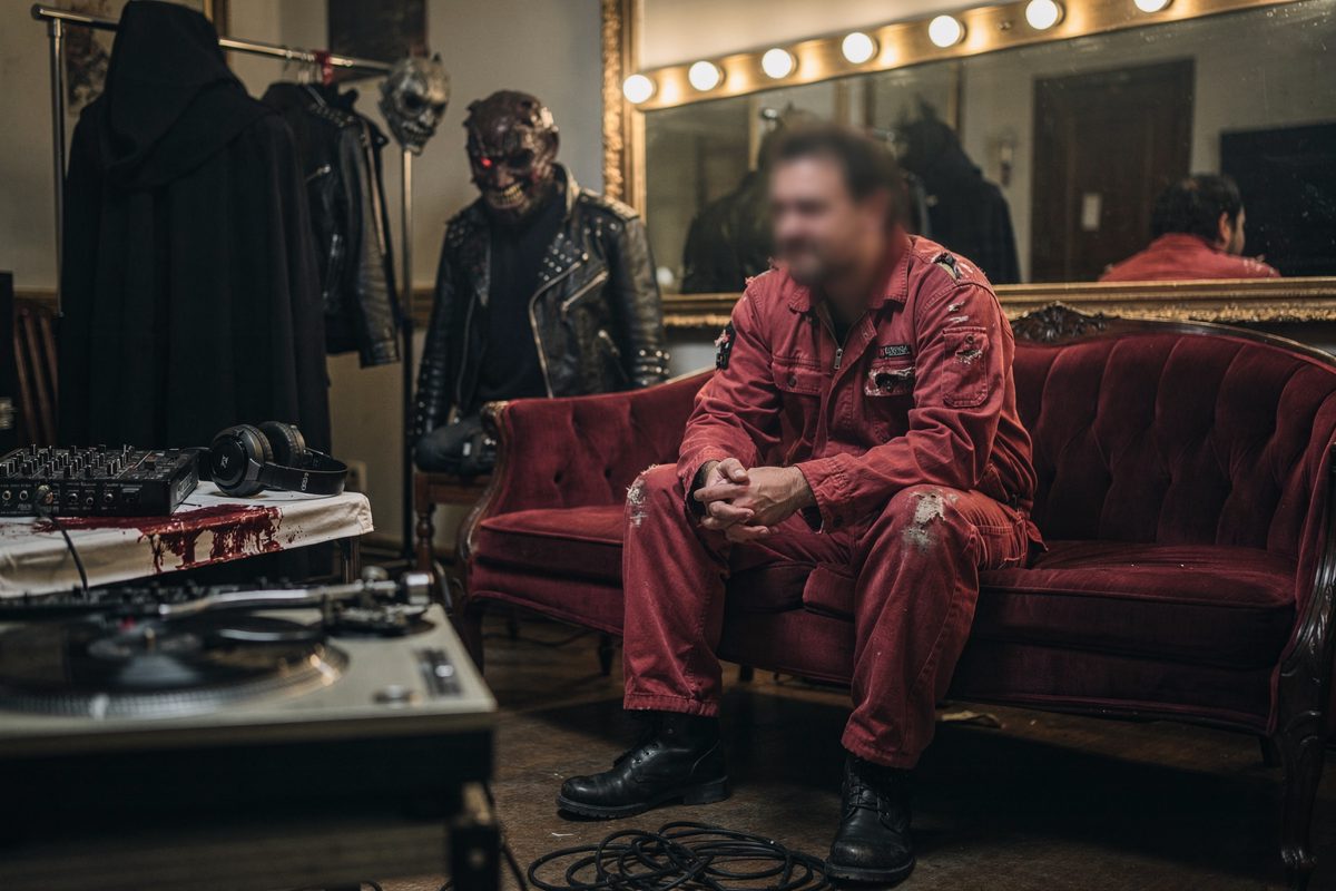A blurred figure in a distressed red industrial boiler suit sits alone on a velvet chaise lounge in a dimly lit, opulent dressing room.