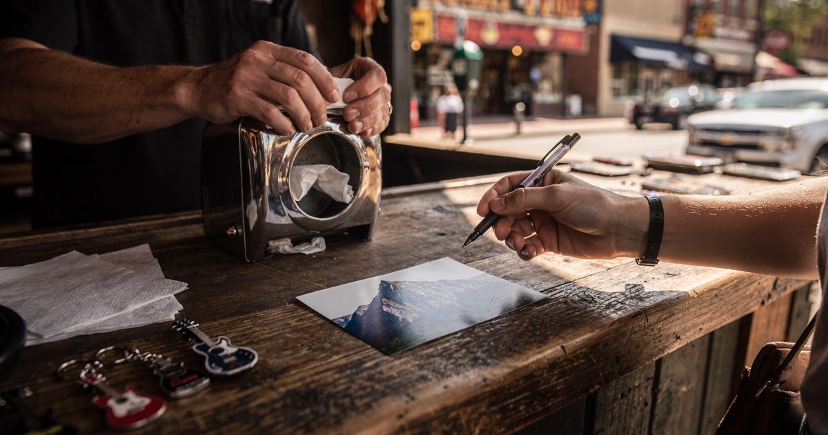 Close shot in hard late-morning sun on a honky-tonk district souvenir counter, a clerk's hands tipping a metal napkin dispenser upside down over the wood while.