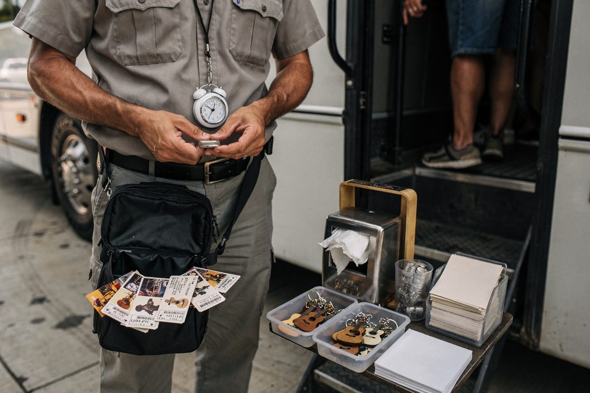 Overhead morning light inside a tour-bus boarding bay, a driver's hands checking a kitchen timer clipped to a lanyard while ticket stubs fan from an open pouch.