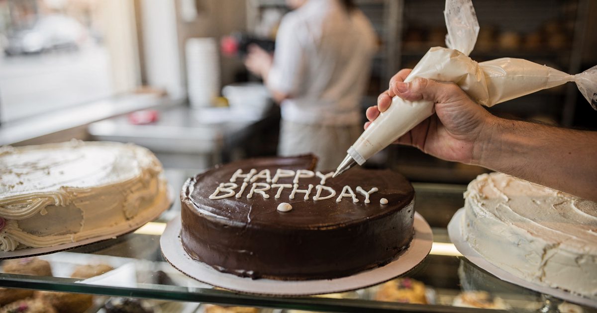 Close detail at a bakery counter in bright mid-morning window light, a baker's hand cropped at the wrist squeezing a piping bag to finish white icing on a.