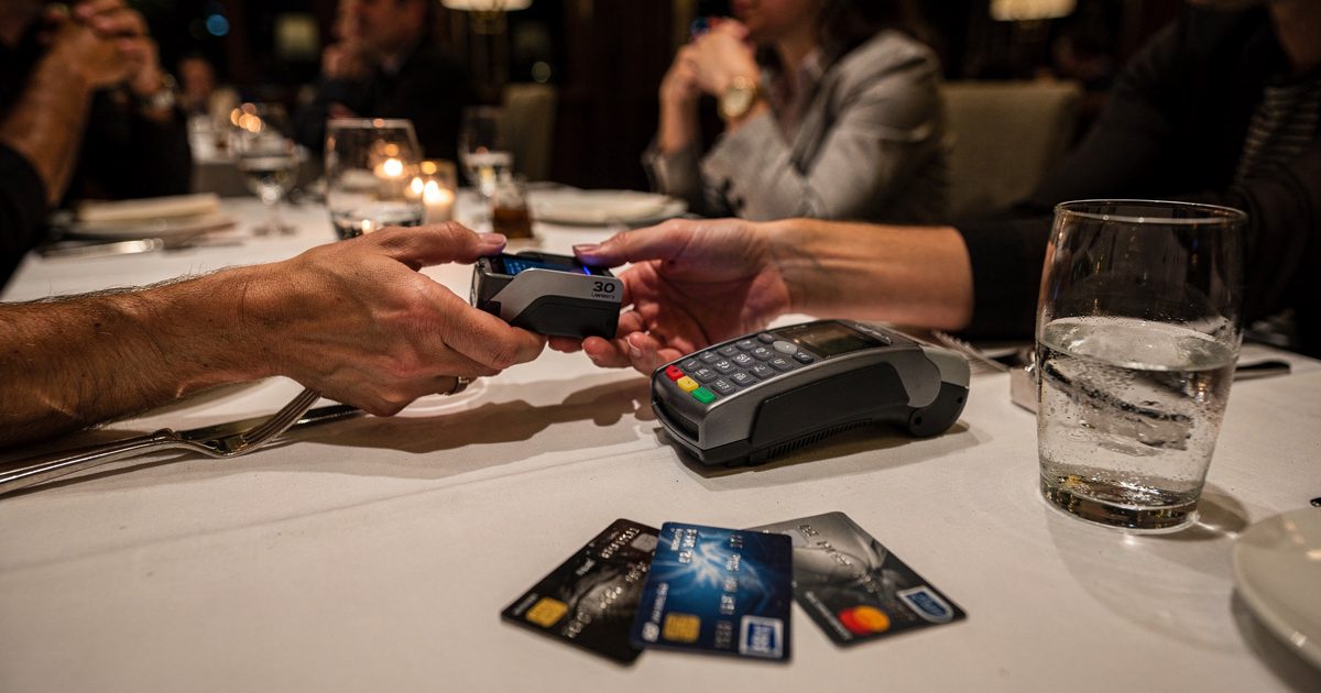 Close-up of two hands passing a folded card machine across a white tablecloth in a dimly lit London restaurant at night, three bank cards fanned beside a.