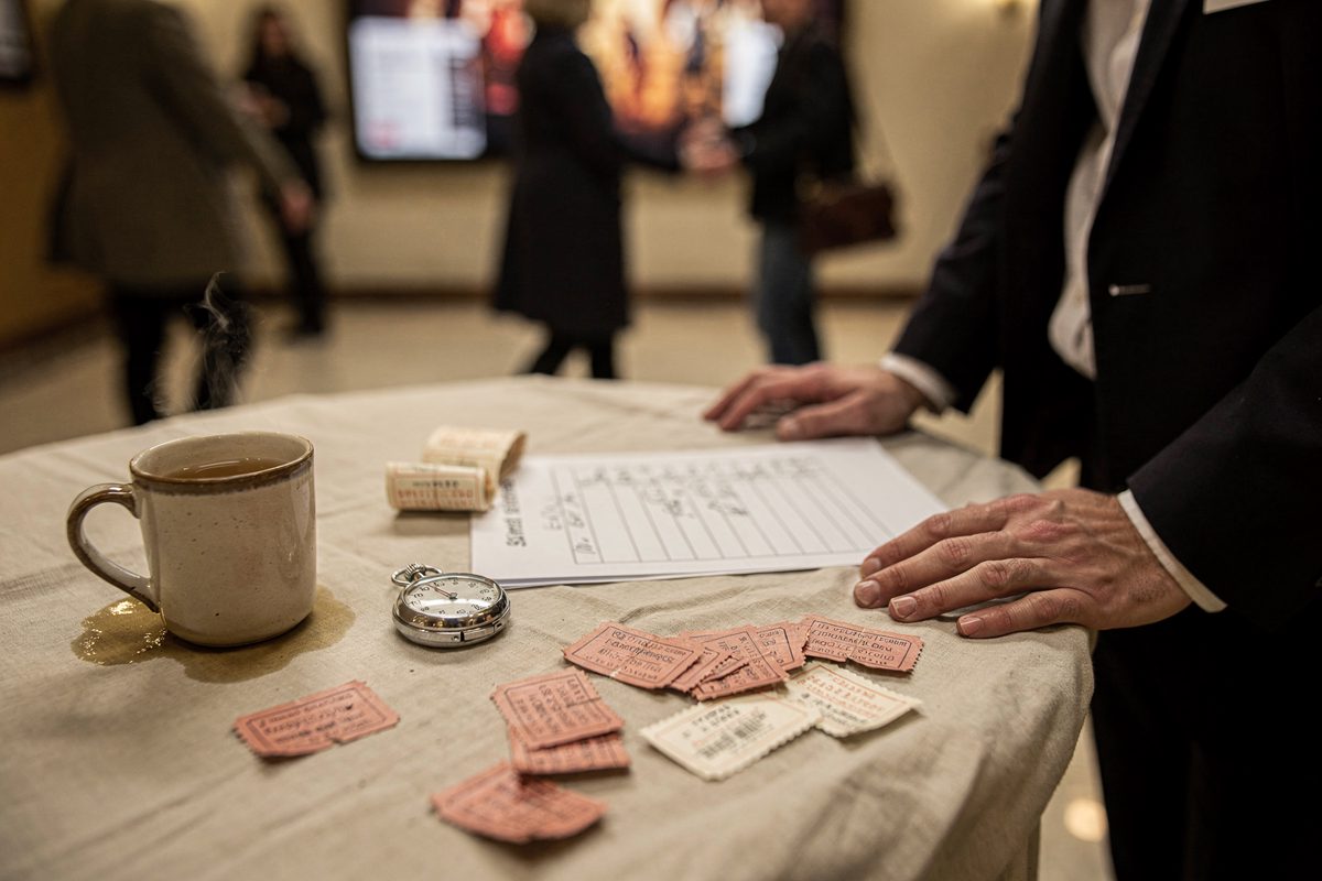 Close mid-morning detail of a foyer folding table where an usher's hands hold a stopwatch beside a handwritten shift rota and a cooling mug of tea, ticket.