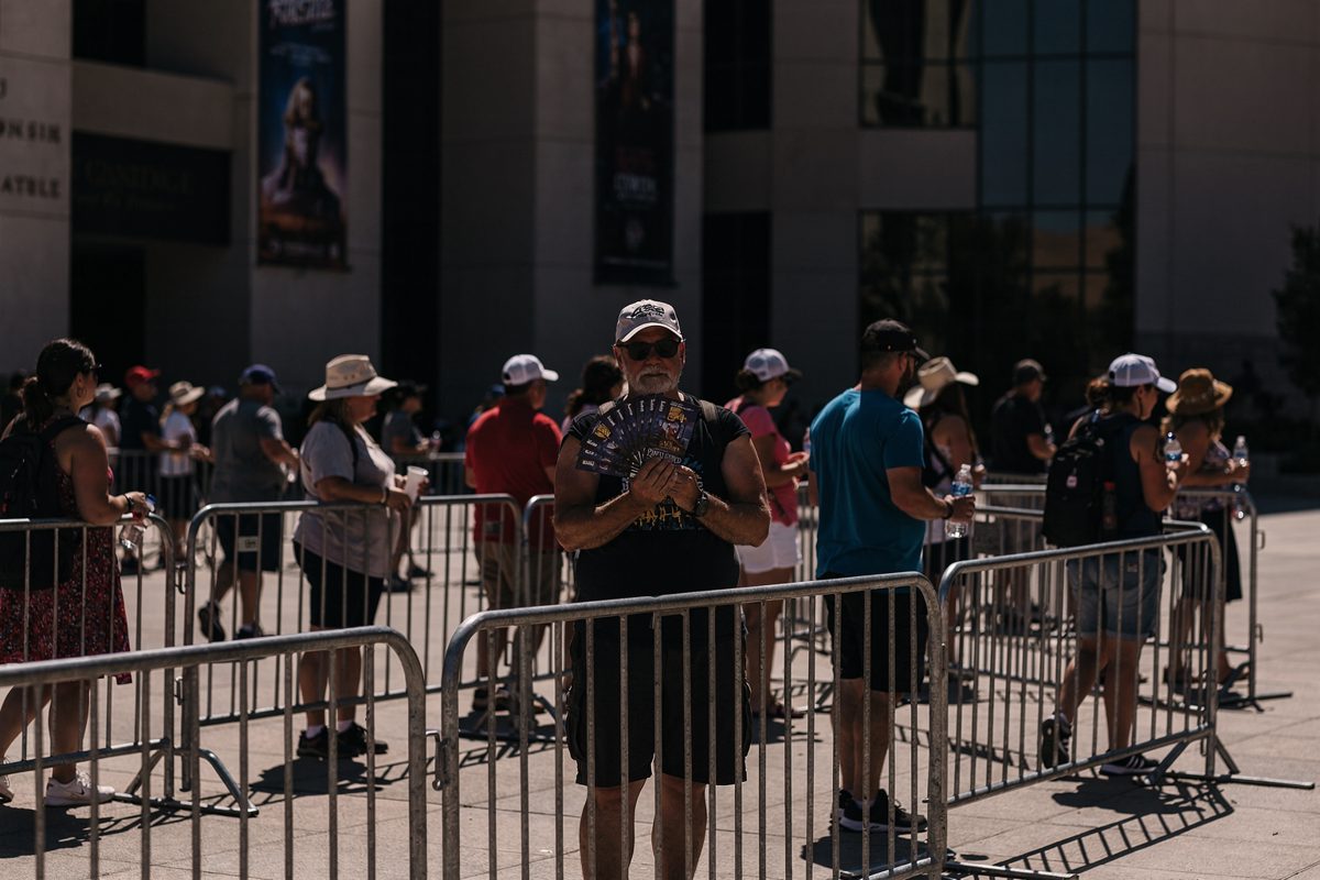 Harsh midday desert sun in a wide shot of tourists silhouetted along crowd barriers snaking across a concrete courthouse plaza, one fanning themselves with a.
