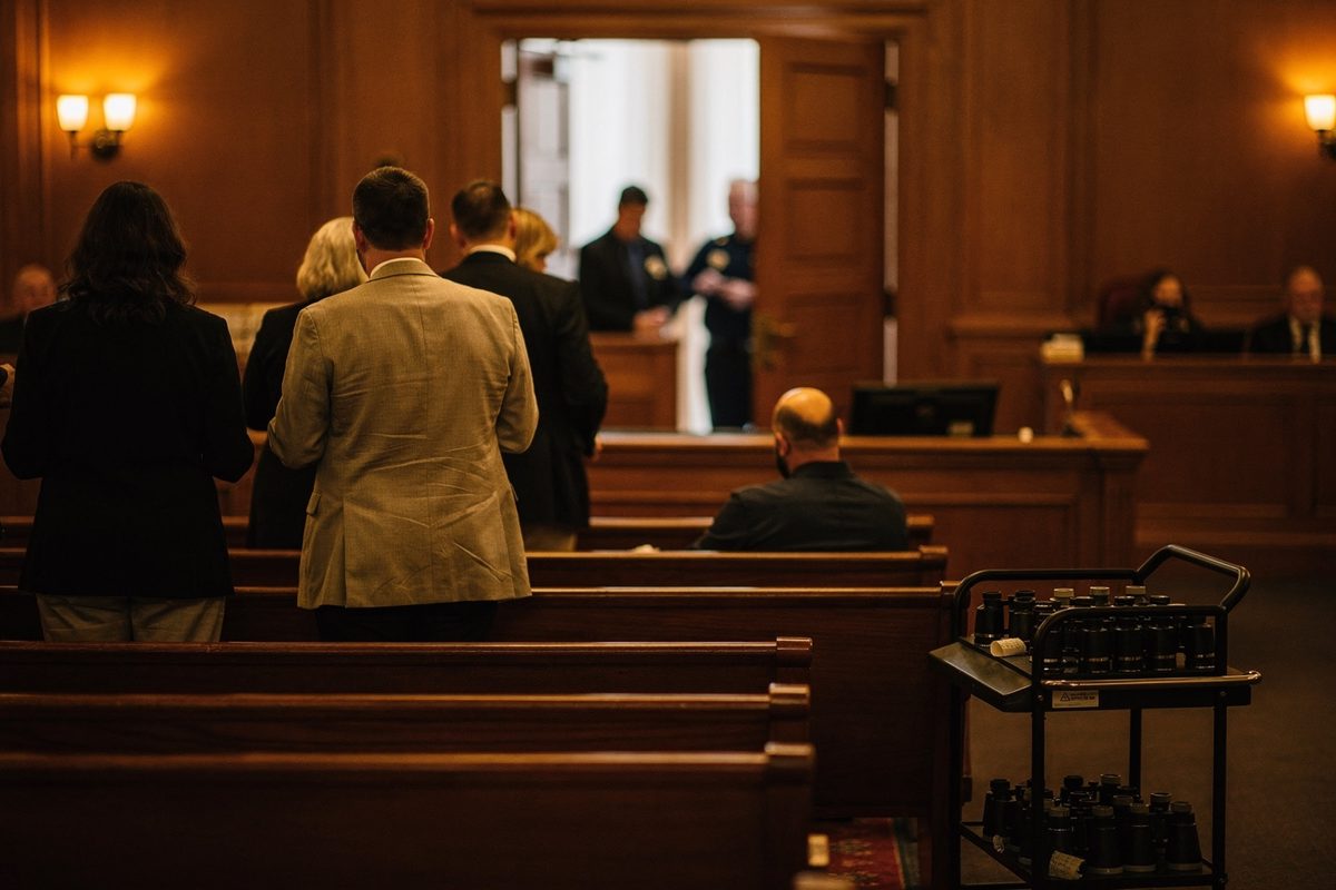 Warm tungsten house light in a wood-panelled courtroom gallery as an usher, backlit to a silhouette at the double doors, tears tickets for a filing line of.
