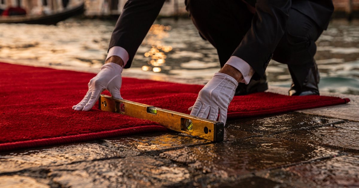 Close detail in low golden evening light on a Venice hotel water landing as two pairs of white-gloved hands ease a crimson carpet flat against wet stone, a.