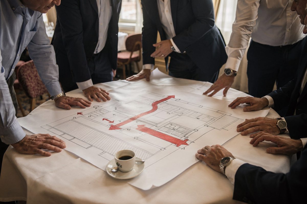 Mid-morning light through tall windows of a Lido hotel dining room, a cluster of officials cropped at the shoulders leaning over a hand-drawn floor plan spread.