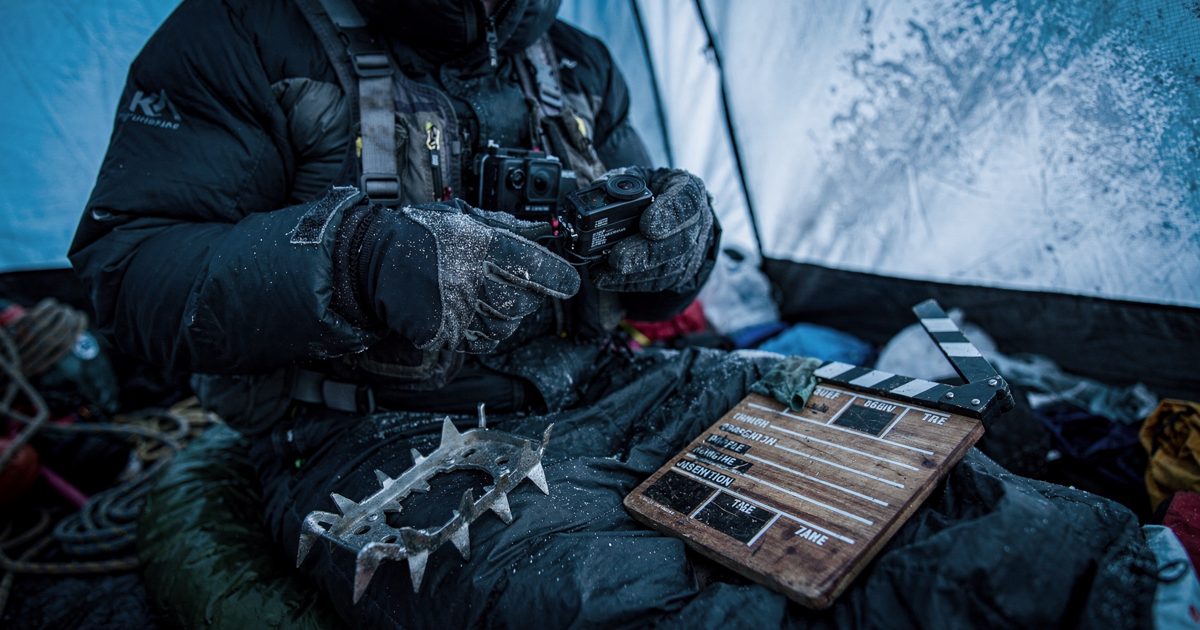 Close shot inside a high-altitude base camp tent in cold blue predawn light, a climber's gloved hands cropped at the cuffs clipping a small action camera to a.