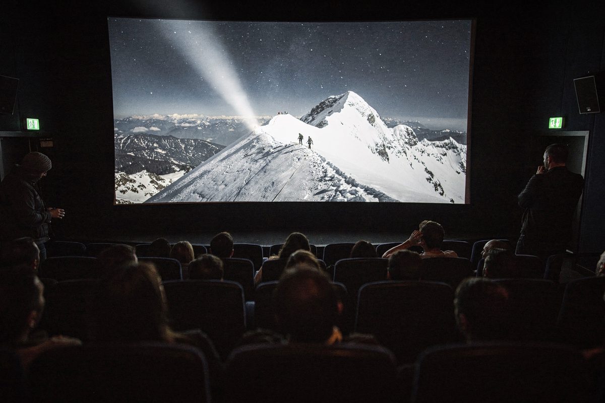 Wide shot inside a dim festival screening room at night, the audience reduced to dark silhouettes against a screen filled with a white snow ridge, a projector.