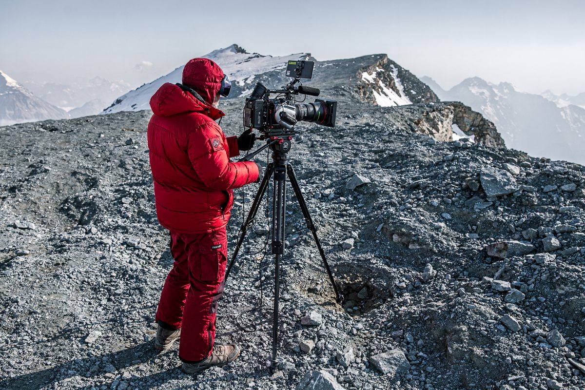 Medium shot on a glacier moraine in flat harsh midday light, a camera operator in a red down suit seen from behind panning a long lens across an empty rocky.