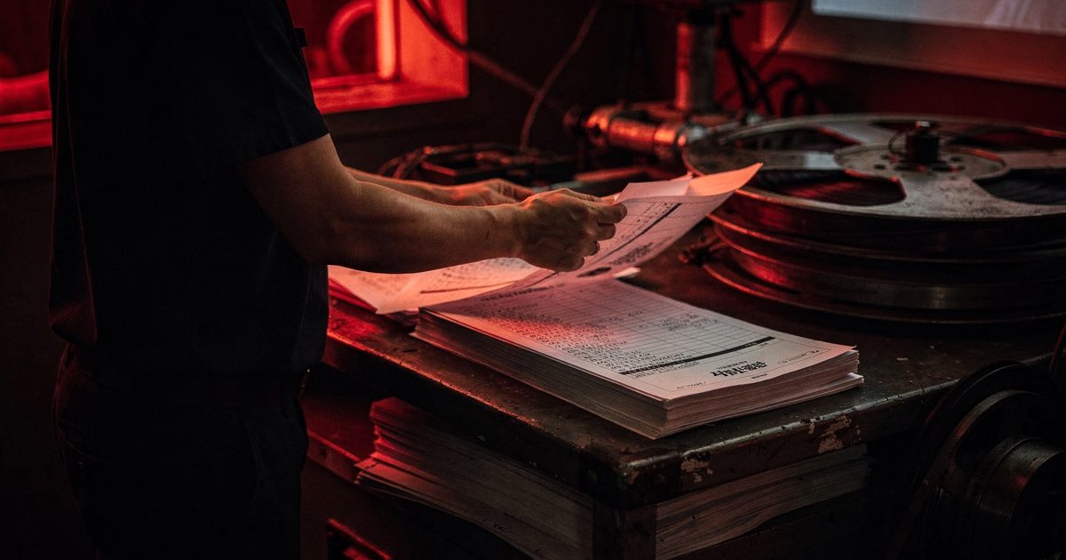 Close detail in the dim red glow of a cinema projection booth at night, a technician's hands fanning out a stack of printed running-order sheets beside a reel.