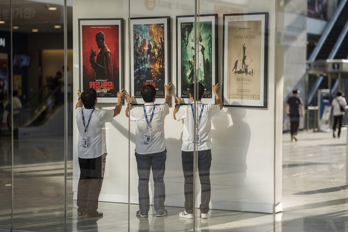 Mid shot in bright morning light through the glass frontage of a Busan multiplex foyer, three festival staff in lanyards seen from behind rearranging framed.