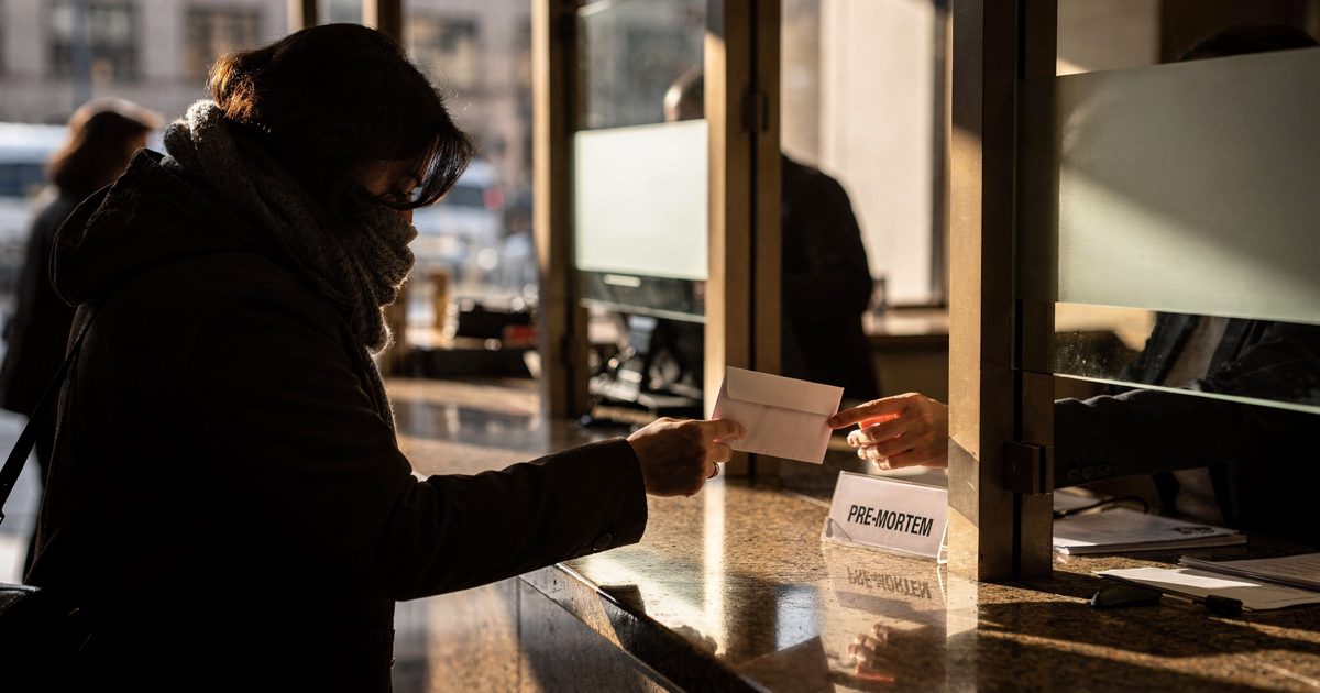 In bright low Saturday sun slanting through the glass of a downtown bank branch, a customer with their face lowered into a scarf extends an envelope across a.