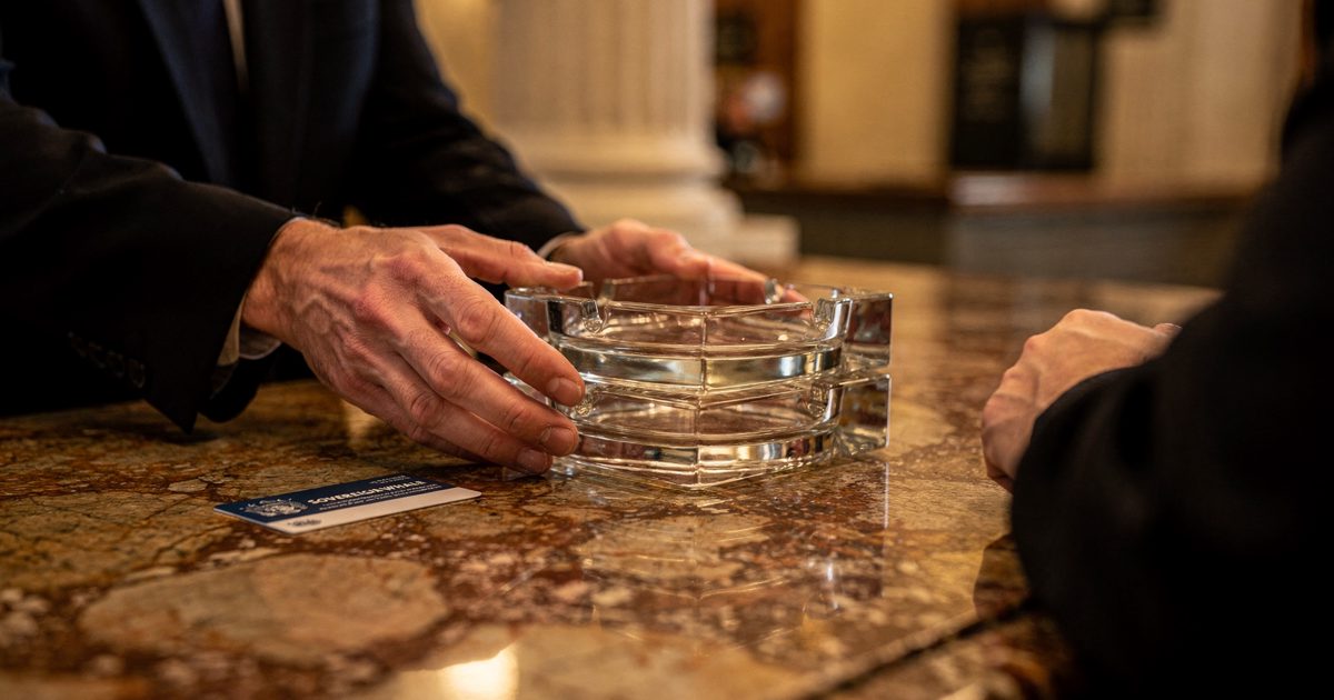 Close detail in warm lamplight on a marble government lobby counter, hands cropped at the wrists sliding a stack of glass ashtrays toward a visitor beside a.