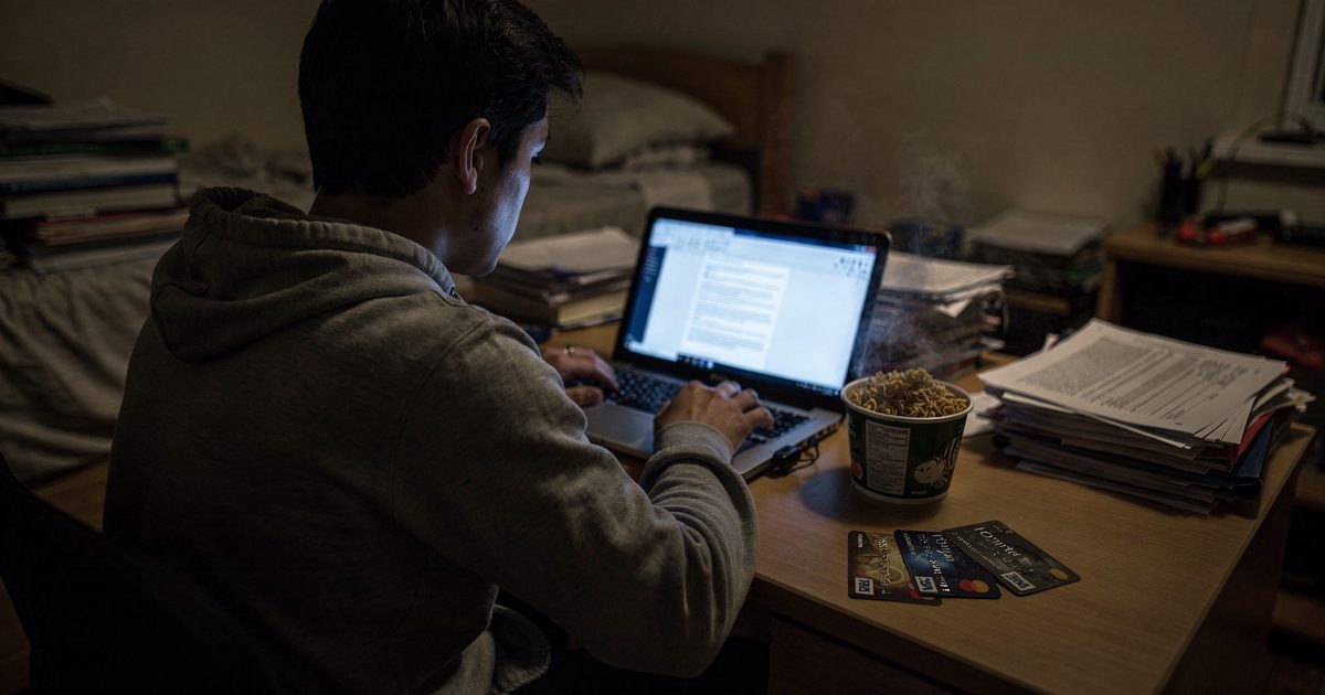 Medium shot inside a cramped dorm room late at night lit only by a laptop screen, a student seen from behind in a hoodie hunched at a desk, three credit cards.