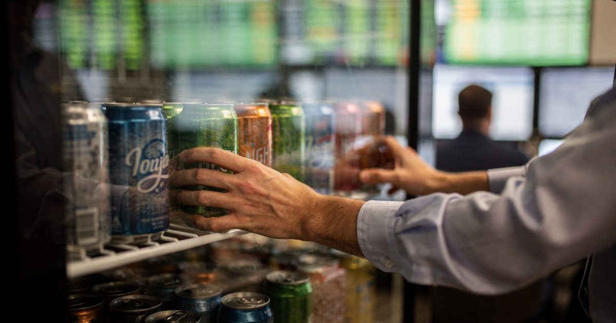 Close crop of a clerk's hands on a Houston energy trading floor in late-afternoon window light, sliding cans of sparkling water onto a glass fridge shelf while.