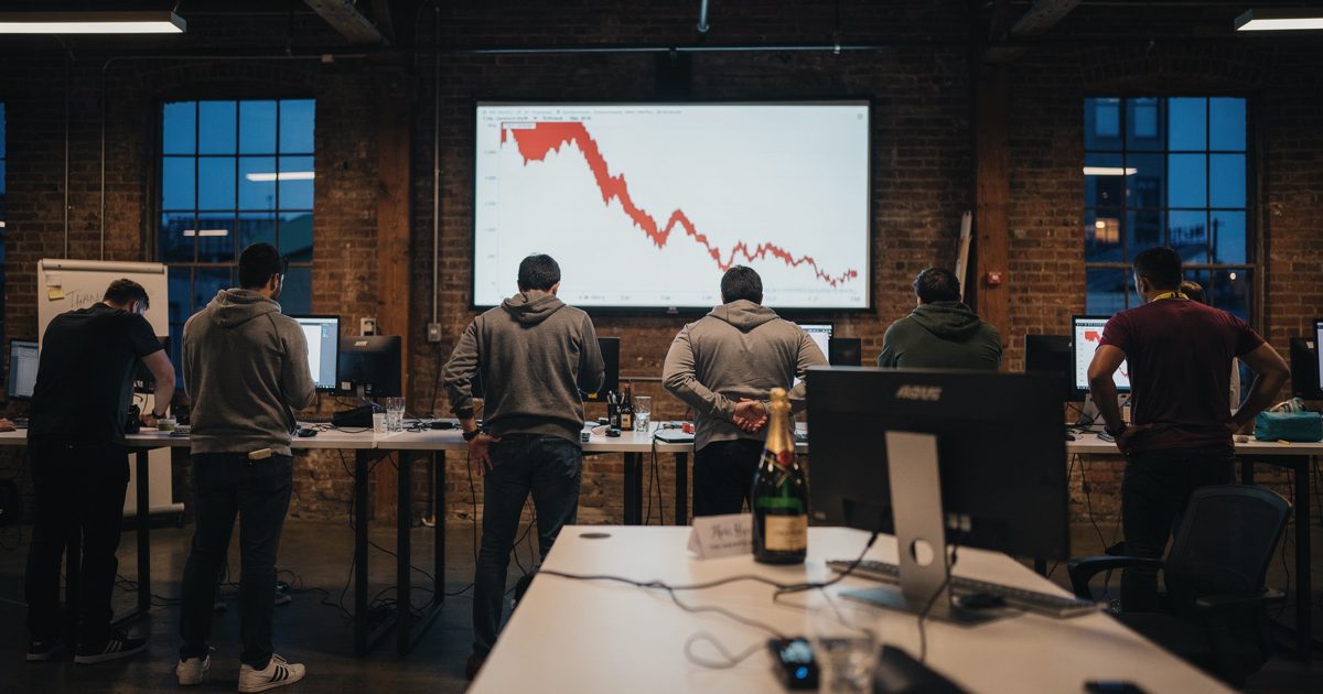 Wide low-angle shot at dusk inside a brick warehouse-loft fintech office in San Francisco, a dozen engineers at standing desks facing away toward a wall-sized.