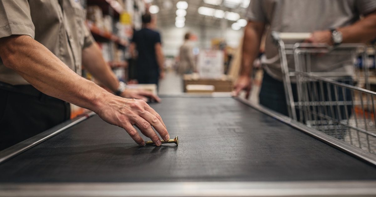 Mid-morning light through high warehouse skylights in a big-box home store, a cashier's hands sliding one brass wood screw across a black checkout belt as a.