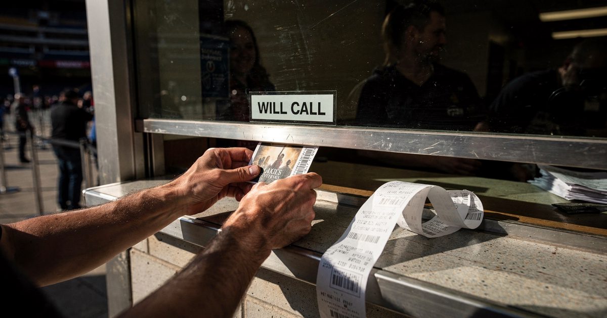 Close detail in harsh late-afternoon sun at an arena box office, a pair of hands cropped at the wrists sliding a thermal-printed ticket through the gap beneath.