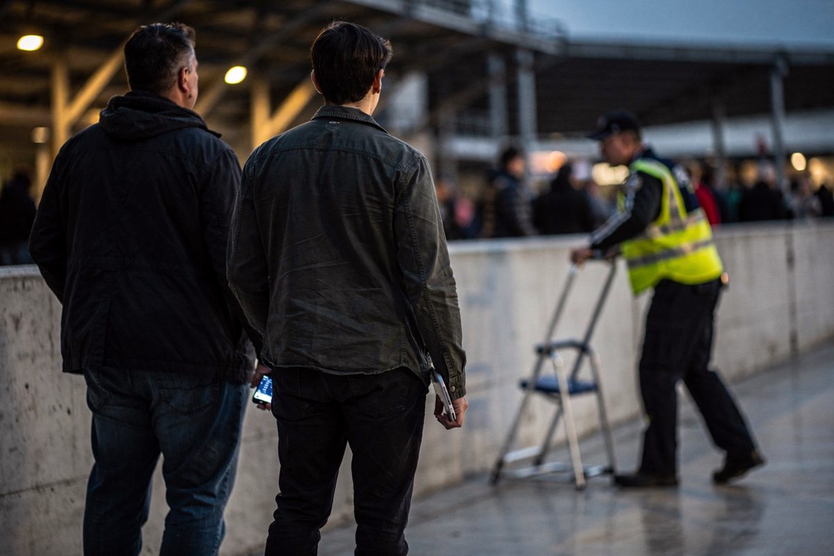 Mid-shot at dusk on an arena concourse, three people seen from behind in a slow queue along a painted concrete wall while a staffer in a high-visibility vest.