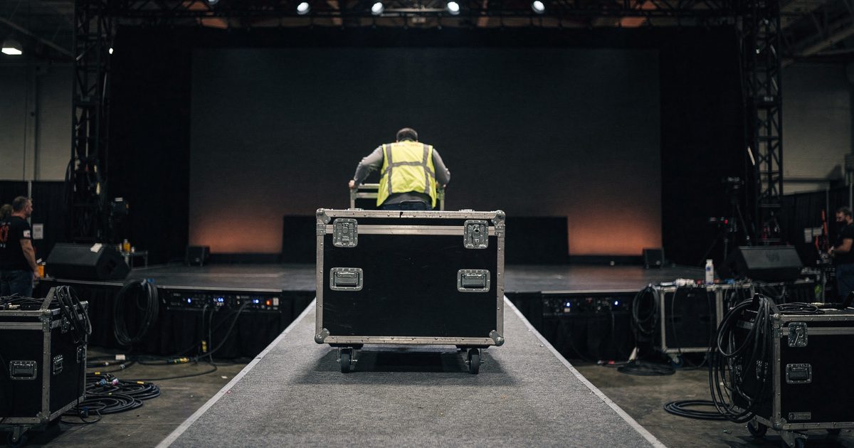 Flat overhead lighting in a cavernous convention center hall, a production worker seen from behind pushing a heavy black flight case up a temporary carpeted.