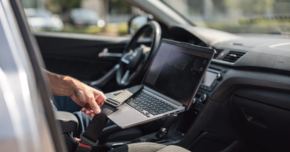Close detail through the open driver's door of a parked sedan in bright mid-morning sun, an open laptop propped on the passenger seat with the seatbelt pulled.