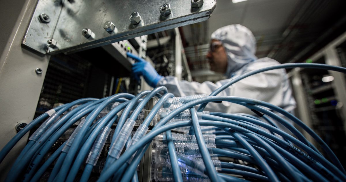 A close upward shot of a tangled bundle of thick, blue fiber-optic server cables pulsing with harsh white light as an out-of-focus technician in a sterile suit.