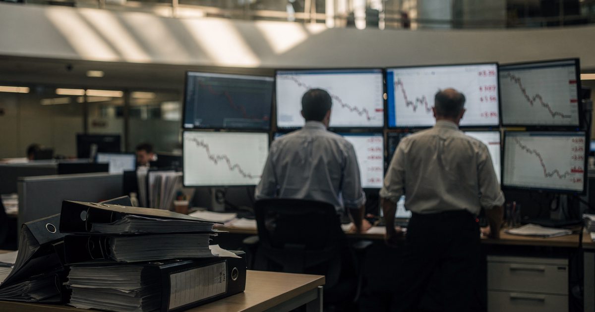 Late-afternoon sun slants from an upper walkway onto an open trading floor where three figures in shirtsleeves, seen only from behind, stand at terminals with.