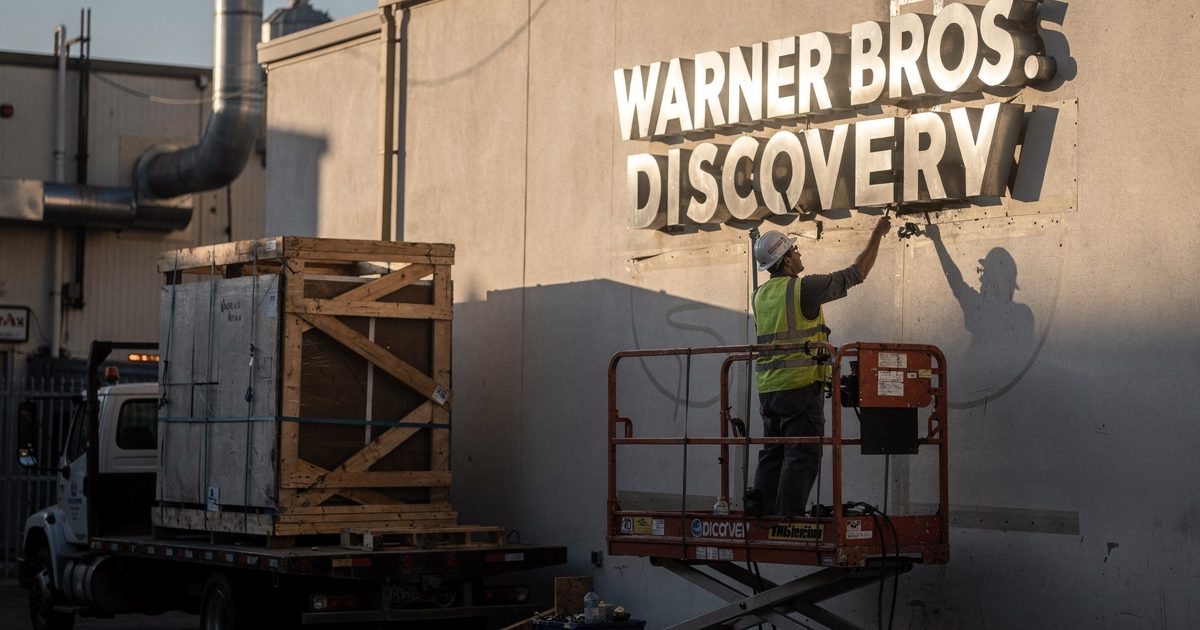 Medium shot from below in slanting late-afternoon sun on a Hollywood studio backlot, a worker in a hard hat seen from behind on a scissor lift unbolting a.