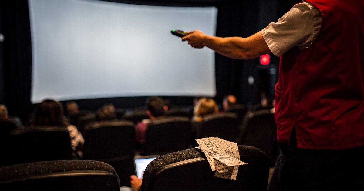 Mid-shot in dim pre-show auditorium light inside a suburban multiplex, an usher in a red vest seen from behind holding a television remote up toward the blank.