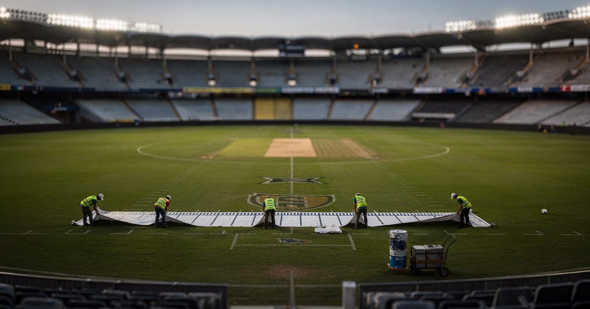 Pale first light over an Australian stadium bowl shot from high in the empty upper tier, a groundcrew of six seen from above laying a long yard-line stencil.