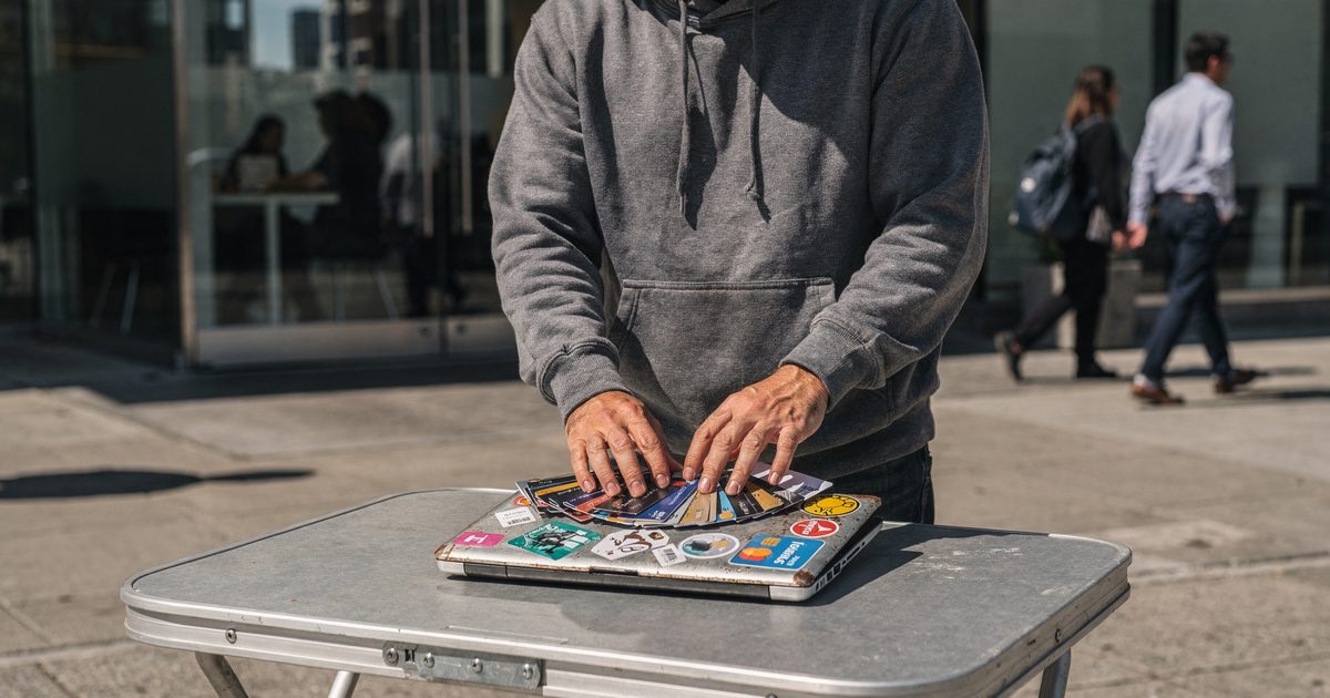 Mid shot in hard midday sun on a SoMa sidewalk outside a glass office lobby, a figure in a grey hoodie cropped at the shoulders fanning a full hand of credit.