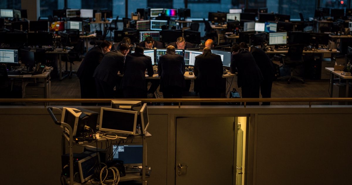 High angle at dusk from a mezzanine rail above a Wall Street trading floor, a tight cluster of fourteen staff in dark suits gathered at a single lit post.