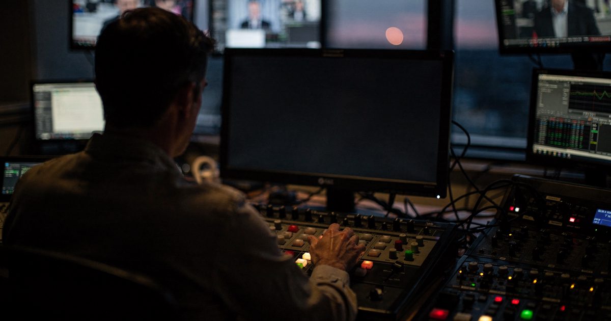 Dim ambient light inside a television control room at dawn, a technician framed from behind reaching toward a complex mixing console while the main preview.